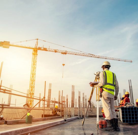 Kỹ sư đội mũ bảo hộ và áo phản quang đo đạc bằng máy trắc địa trên công trường, phía sau là cần cẩu tháp và các công nhân