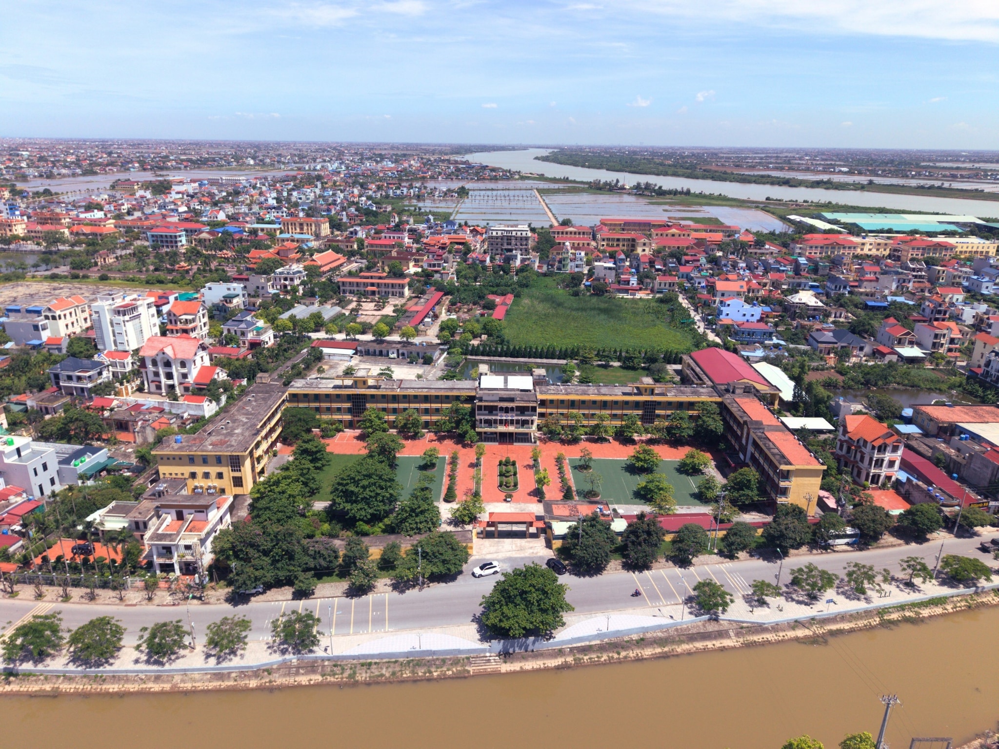 Ảnh flycam khu dân cư ven sông ở Ninh Bình: ngôi trường nhiều tầng với sân gạch đỏ, nhà dân mái ngói xung quanh, đường ven sông và dòng sông phù sa phía trước